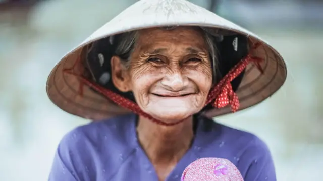 A smiling elderly woman wearing a conical straw hat traditionally associated with field work.
